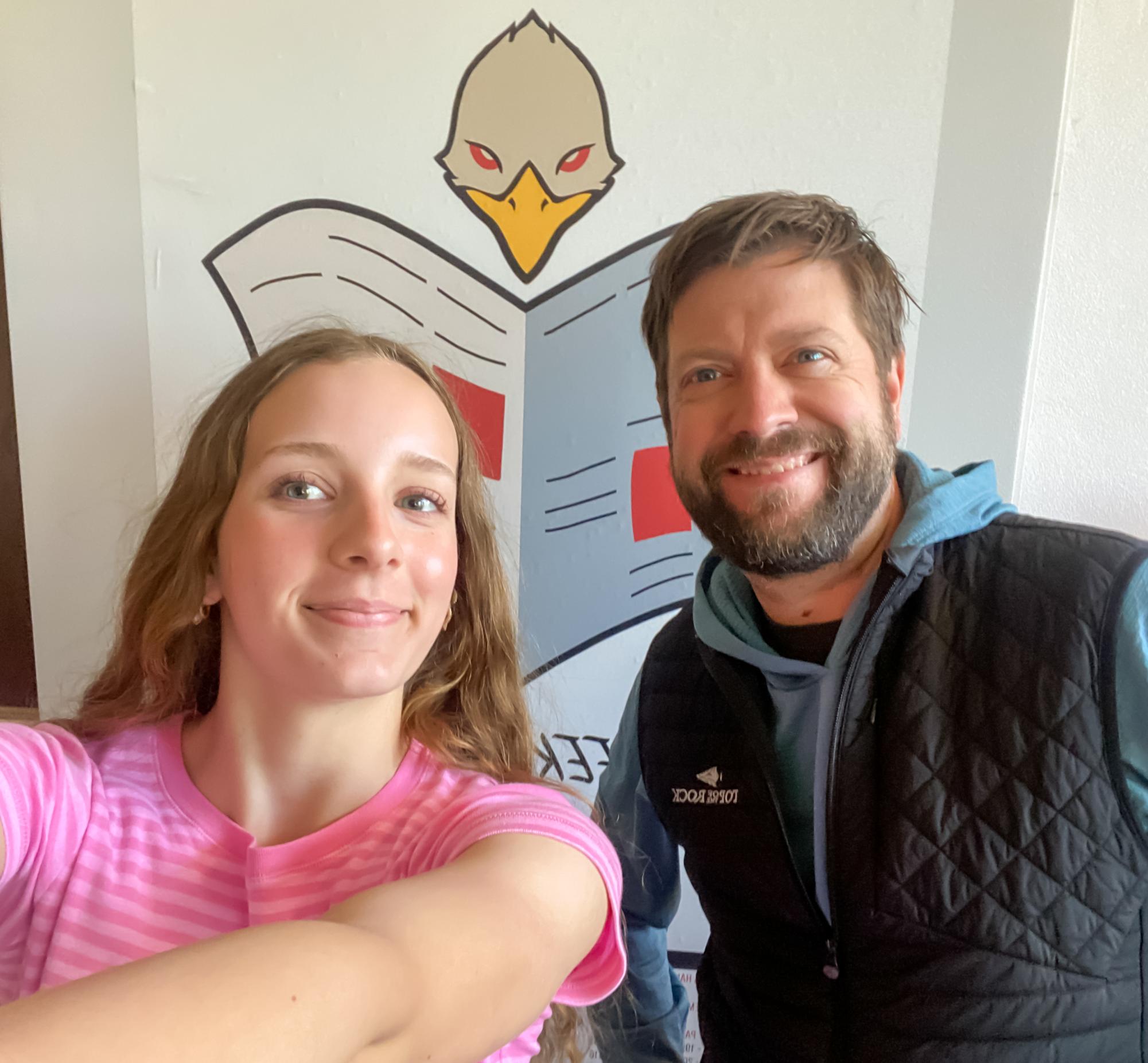 Hazel and Rog pose in front of the Little Hawk journalism logo with the quote "Seek Truth and Report It".